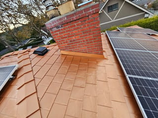 Copper Penny Interlock Slate aluminum roof being installed on a brown single-storey home with a chimney among trees, Victoria, British Columbia