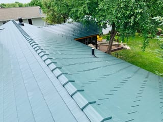 Hunter Green Interlock Shingle aluminum roof on a green two-storey home with dormers among tall trees, Winnebago, Illinois