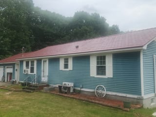 Single-story ranch home with Interlock tile-red aluminum slate metal roof, blue vinyl siding, white shutters, and decorative wagon wheel accents, Lebanon, ME.