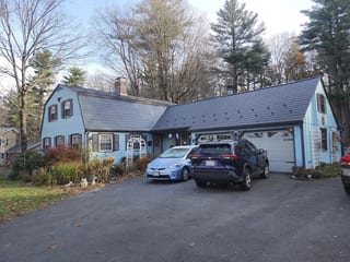 Gambrel-style colonial home with Interlock deep charcoal cedar shingle metal roofing, chimney, and attached garage in a forested Westborough, MA suburb.