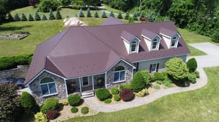 Aerial front view of a 1.5-story Colonial home in NH featuring an Interlock Colonial Brown cedar shingle metal roof with dormers and stone facade.