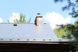Deep charcoal Interlock aluminum slate metal roof on a rural NH home, featuring a wood-clad chimney with cap, roof louver, and decorative eave details.