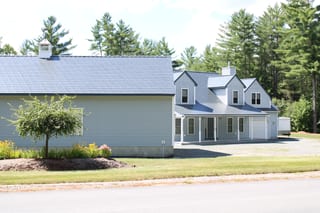 Two-story farmhouse in Thornton NH with Interlock deep charcoal aluminum slate metal roofing, multiple dormers, covered porch, and attached garage amid tall pines.