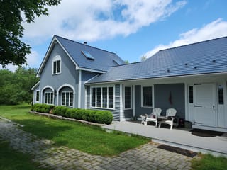 Front-left view of a blue-gray New Hampshire home with Interlock deep charcoal aluminum slate roof, skylights, dormers, and covered porch with Adirondack chairs.