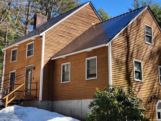 Two-story colonial home with black Interlock aluminum slate metal roofing, warm cedar lap siding, and a brick chimney set in a forested New Hampshire winter landscape.