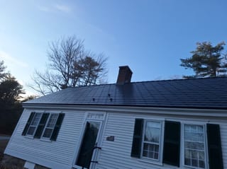 White Colonial ranch home in Hillsboro NH with Interlock deep charcoal aluminum slate roof, central brick chimney, and green shutters under a clear blue sky.