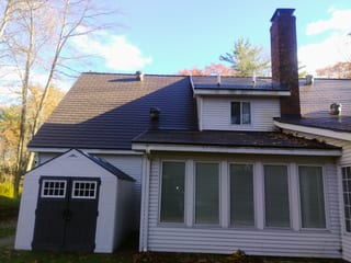 Tudor Brown Interlock aluminum cedar shingle metal roof on a 2-story colonial home with dormer, brick chimney, and multiple roof planes, fall foliage backdrop.