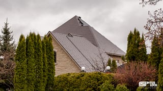 Large Tudor-style stone home in Sudbury ON featuring a steeply pitched multi-hip Interlock aluminum slate roof in Tudor Brown, surrounded by mature evergreens.