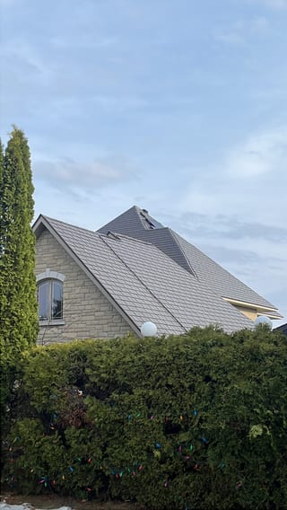 Steep multi-hip Interlock aluminum slate roof in Tudor Brown on a stone two-storey home in Sudbury, ON, framed by tall cedars and holiday lights.