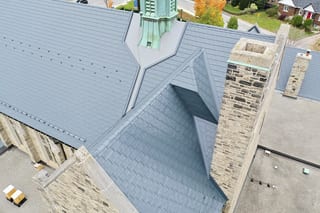 Aerial view of a deep charcoal Interlock aluminum slate roof on a historic stone church in Toronto, featuring complex valleys, a patinated copper spire, and stone chimneys.