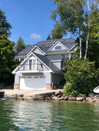 Craftsman lakefront home with deep charcoal Interlock aluminum slate roof, multiple solar panels, complex multi-gable profile, and boathouse garage on rocky shoreline.