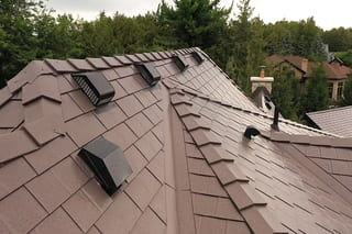 Complex Tudor Brown Interlock aluminum slate metal roof with multiple valleys, roof louvers, vent pipes, and ridge caps on a suburban forested home.
