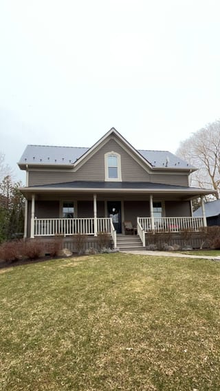 Two-storey farmhouse in Ravenna, ON with black Interlock aluminum slate metal roof, front dormer, wrap-around porch, and snow guards visible on steep gable.