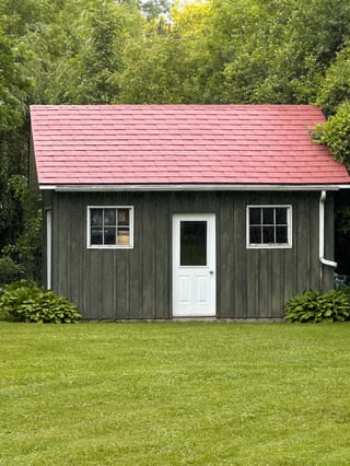 Front elevation of a rustic wood-clad outbuilding with a Tile Red Interlock aluminum shingle gable roof, white door, and twin windows in Almonte, ON.