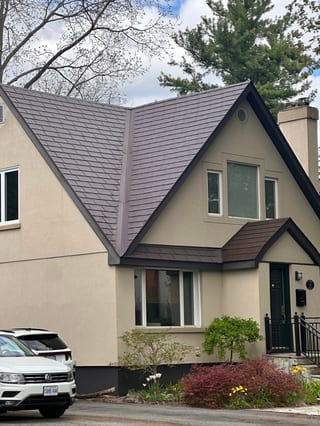 Two-storey Tudor-style home with a complex steep-pitched Colonial Brown Interlock aluminum slate roof featuring dormers, valleys, and a chimney.