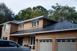 Two-storey brick colonial home with deep charcoal Interlock aluminum slate metal roof, snow guards visible, set against a forested suburban backdrop.
