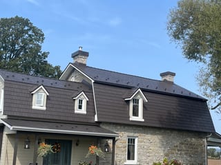 Tudor Brown Interlock aluminum cedar shingle metal roof on a 1.5-storey heritage stone home in Rockland, ON, featuring three dormers and two stone chimneys.