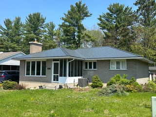 Single-storey ranch-style brick home with a deep charcoal Interlock aluminum slate hip roof, chimney, and screened front entry, set against tall pines.