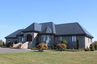 Large stone estate home in Alexandria ON with a complex deep charcoal Interlock aluminum slate roof featuring a central turret, steep hip planes, and manicured topiary landscaping.