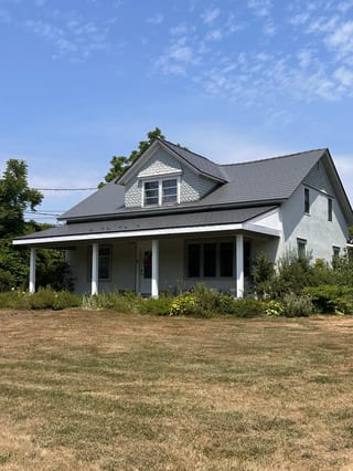 Musket Grey Interlock Slate aluminum roof on a grey two-storey home with dormers among tall pines, Bloomfield, Ontario