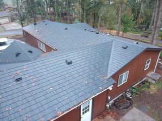 Aerial view of a complex multi-hip Interlock aluminum slate roof in charcoal black on a red-sided home surrounded by tall firs in Albany, OR.