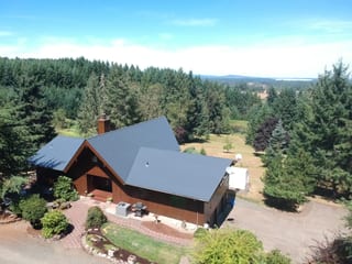 Aerial view of a chalet-style home in Eugene, OR with a deep charcoal Interlock cedar shingle metal roof, prominent gable, and chimney surrounded by dense forest.