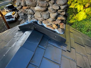 Close-up of black Interlock aluminum slate roof panels with custom metal step flashing around a river-rock stone chimney, Alsea OR.