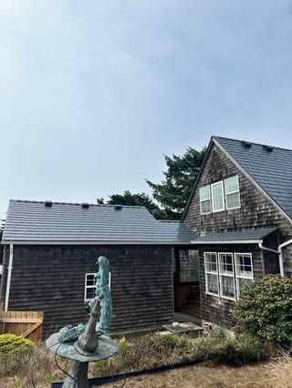 Weathered cedar-shingle coastal cottage in Newport OR with a steep multi-plane Deep Charcoal Interlock aluminum slate metal roof and bronze garden sculpture.