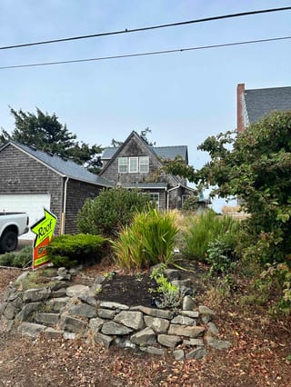 Coastal shingle-style cottage in Newport OR with deep charcoal Interlock aluminum slate roof visible above lush front garden and stone raised bed.