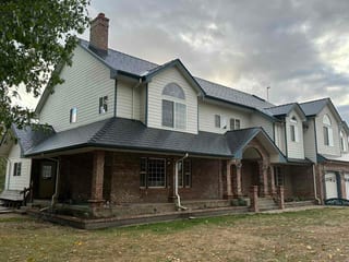 Large two-story Colonial home in Eugene OR with Interlock black aluminum slate metal roofing, brick lower exterior, arched windows, and brick chimney under overcast skies.