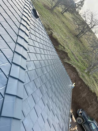 Steep deep charcoal Interlock aluminum slate roof on a rural home in The Dalles, OR, with tightly laid dark metal slate tiles and a hillside backdrop.
