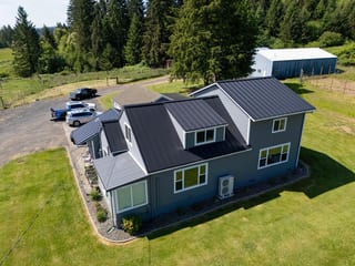 Black Interlock Standing Seam aluminum roof on a dark grey two-storey home with dormers among tall pines, Warren, Oregon
