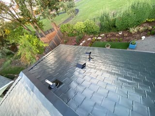 Aerial rear view of a black Interlock aluminum slate metal roof with vent pipes and louvers on a suburban Portland home surrounded by lush landscaping.