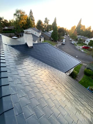 Aerial view of a two-story suburban home with a complex black Interlock aluminum slate metal roof, chimney, and valley details at golden hour in Portland, OR.
