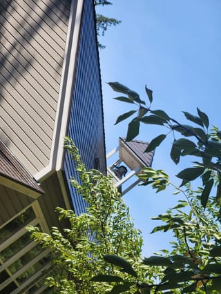 Upward-angle view of a Tudor Brown Interlock cedar shingle metal roof on a multi-gable craftsman home, partially obscured by lush green trees.