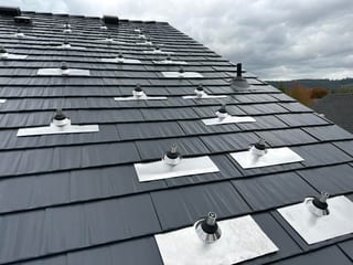 Close-up of deep charcoal Interlock aluminum cedar shingle metal roof with multiple aluminum-flashed vent pipe penetrations on a hip roof.