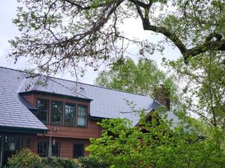 Craftsman home in Corvallis OR with deep charcoal Interlock aluminum slate roof, dormer, stone chimney, and cedar shingle siding framed by mature trees.