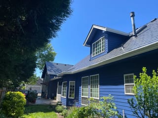 1.5-story Craftsman home with deep charcoal Interlock aluminum slate metal roof, prominent dormer, and vent pipe against a clear blue sky in Canby, OR.