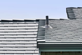 Close-up of Interlock deep charcoal aluminum slate roofing showing multi-level roof transition with vent pipe flashing detail against a clear blue sky.