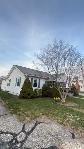 White ranch-style home in Pawtucket RI with Hunter Green Interlock aluminum shingle roof, green shutters, evergreen shrubs, and blooming daffodils.