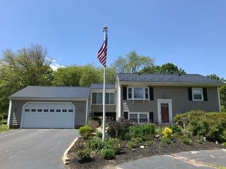 Black Interlock Slate aluminum roof on a grey two-storey home with a metal roof among green trees, Tiverton, Rhode Island