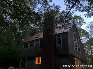 Two-story cedar-shingled modern home with Copper Penny Interlock metal roof, prominent brick chimney, arched window, and skylight at twilight in a forested setting.
