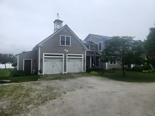 Two-story farmhouse with deep charcoal Interlock aluminum slate roof, cupola with weather vane, double white carriage garage doors, gray shingle siding, rural Vermont setting.
