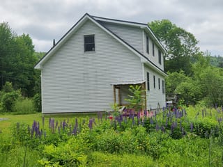 Side elevation of a two-story Vermont farmhouse with Musket Grey standing seam metal roof, surrounded by wild lupines and lush green forest.