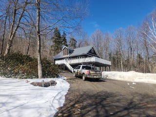 Chalet-style Vermont home with Interlock Musket Grey cedar shingle metal roof, steep gable profile, and white wraparound deck, surrounded by bare winter trees.