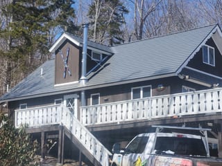 Two-story chalet-style Vermont home with Interlock Musket Grey cedar shingle aluminum roof, front dormer, vent pipe, and white deck railings amid winter forest.