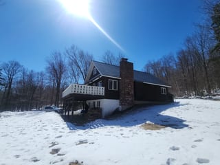 Two-story Vermont chalet with Interlock Musket Grey cedar shingle metal roof, prominent brick chimney, elevated deck, surrounded by winter forest.