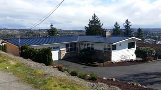Black Interlock Standing Seam aluminum roof on a white ranch-style home with a metal roof among tall trees, Des Moines, Washington