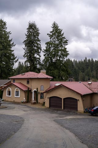 Tile Red Interlock Tile aluminum roof on a tan two-storey home with red roof tiles among tall pines, Woodinville, Washington