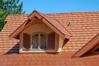 Close-up of a Copper Penny Interlock aluminum shake metal roof on a Craftsman-style home featuring a gabled dormer with arched window and wood shutters.